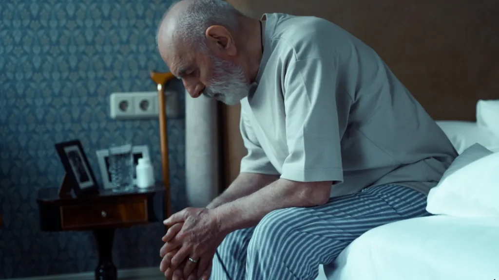 Uma pessoa de joelhos em oração ao lado da cama, com a luz suave do luar entrando pela janela, criando um ambiente sereno e íntimo. Um exemplar aberto da Bíblia está visível no criado-mudo, com uma vela acesa próxima.